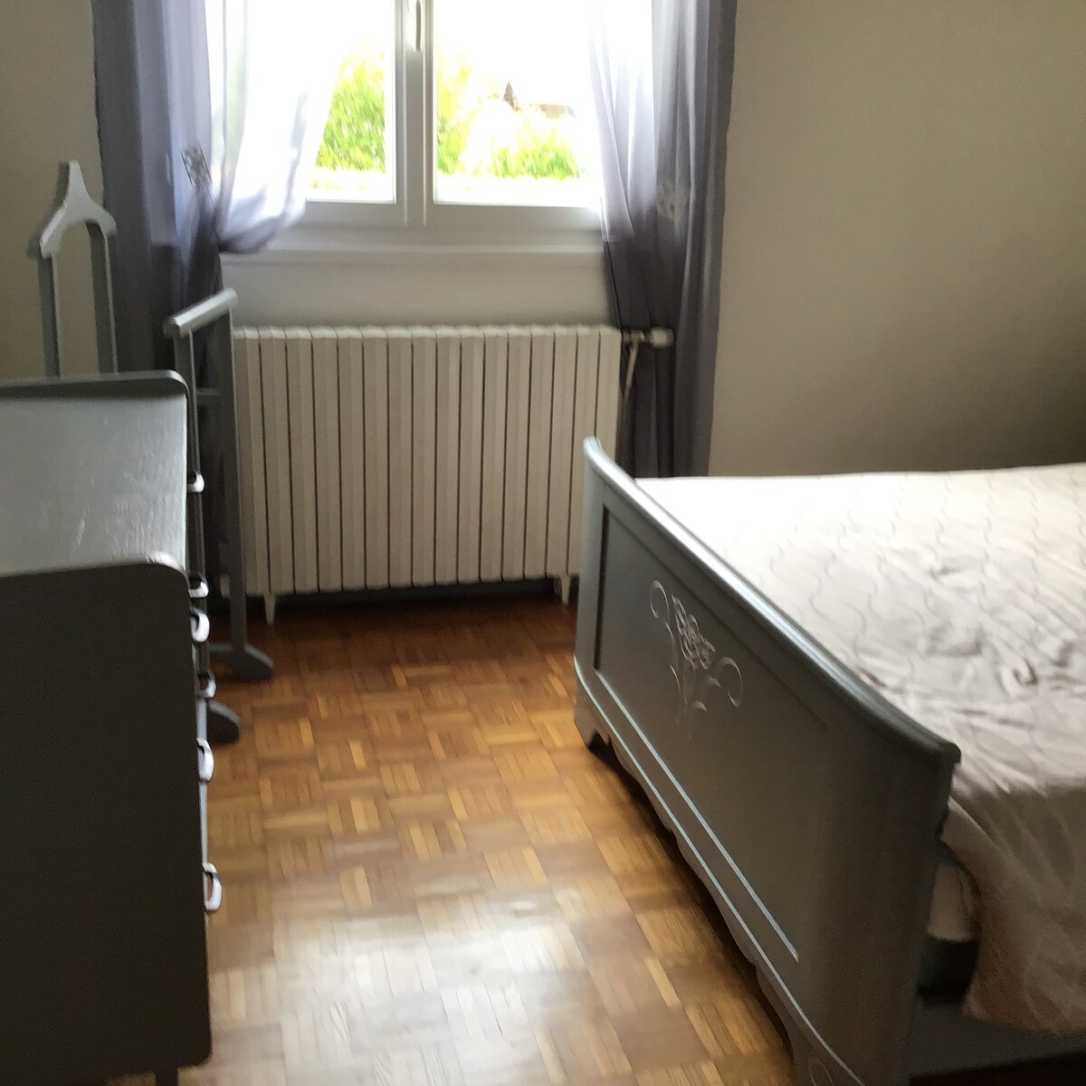 The room features a light-colored wooden floor and a spacious bed with a subtle design. Natural light enters through a window with sheer curtains, illuminating a small dresser and a radiator nearby, enhancing the room's calm atmosphere.