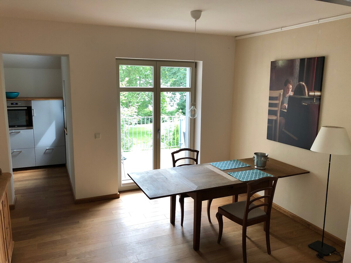 An open dining area features a wooden table with two chairs and a decorative blue table runner. A modern lamp stands beside the table. Natural light streams in through a large window, illuminating a calming view of greenery outside.