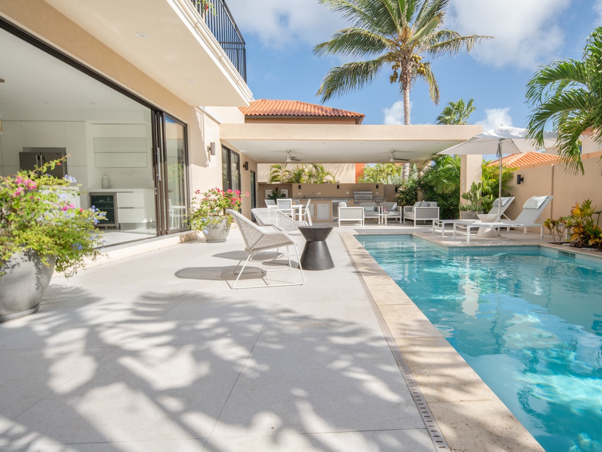 A serene outdoor space showcases a private pool surrounded by lounge chairs and tropical plants. The covered kitchenette is visible in the background, with a dining area adjacent to the pool. Bright sunlight filters through the palm trees, highlighting the villa's spacious layout.