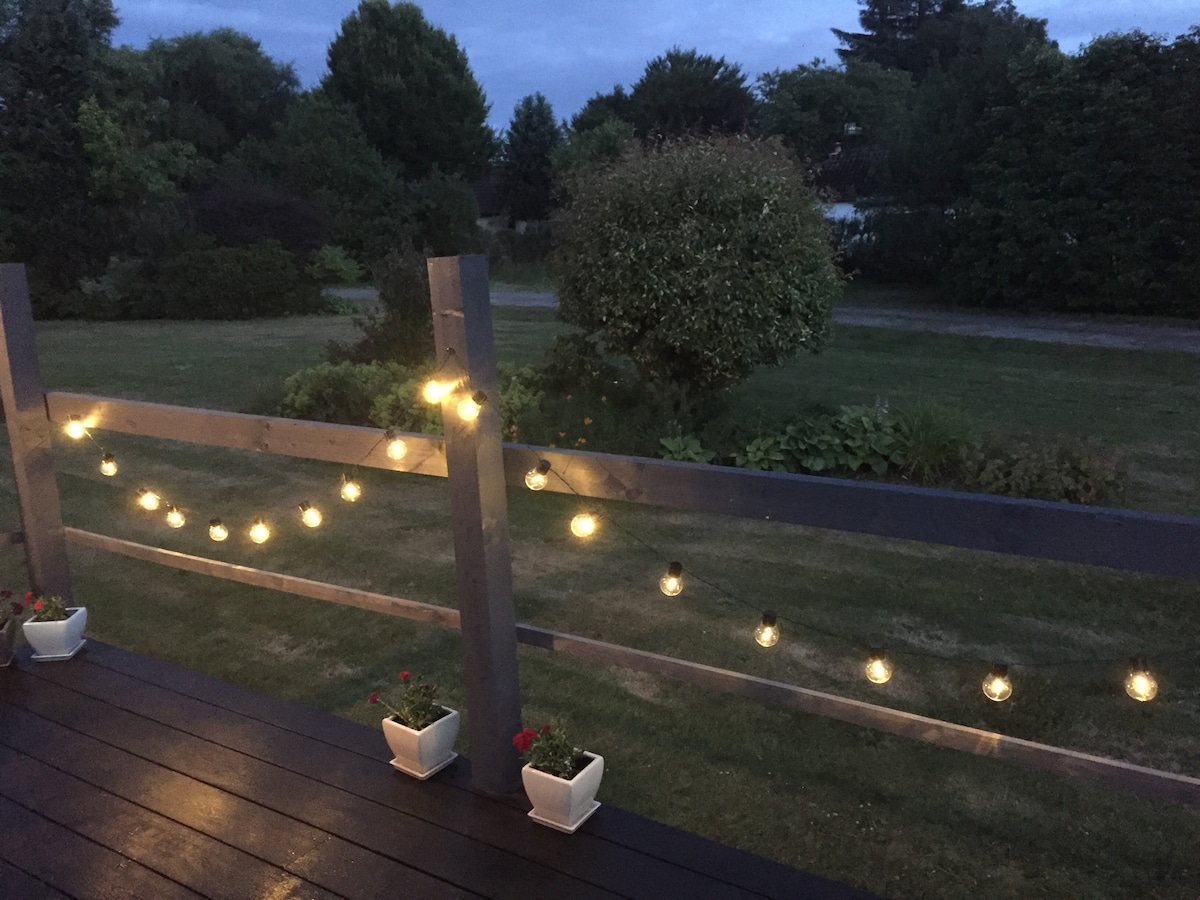 A private decking area is illuminated by string lights, casting a soft glow across the space. Flower pots with plants are placed at the base of the wooden railing, surrounded by a well-maintained grassy area and distant trees, creating a peaceful outdoor setting.