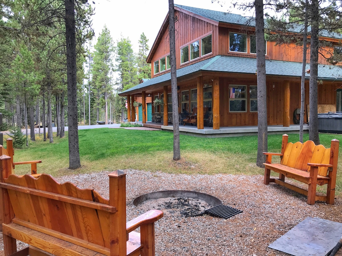 A comfortable outdoor seating area is shown, featuring two wooden benches positioned around a fire pit. A spacious cabin with large windows is visible in the background, surrounded by trees and a grassy lawn, creating an inviting space for gatherings.