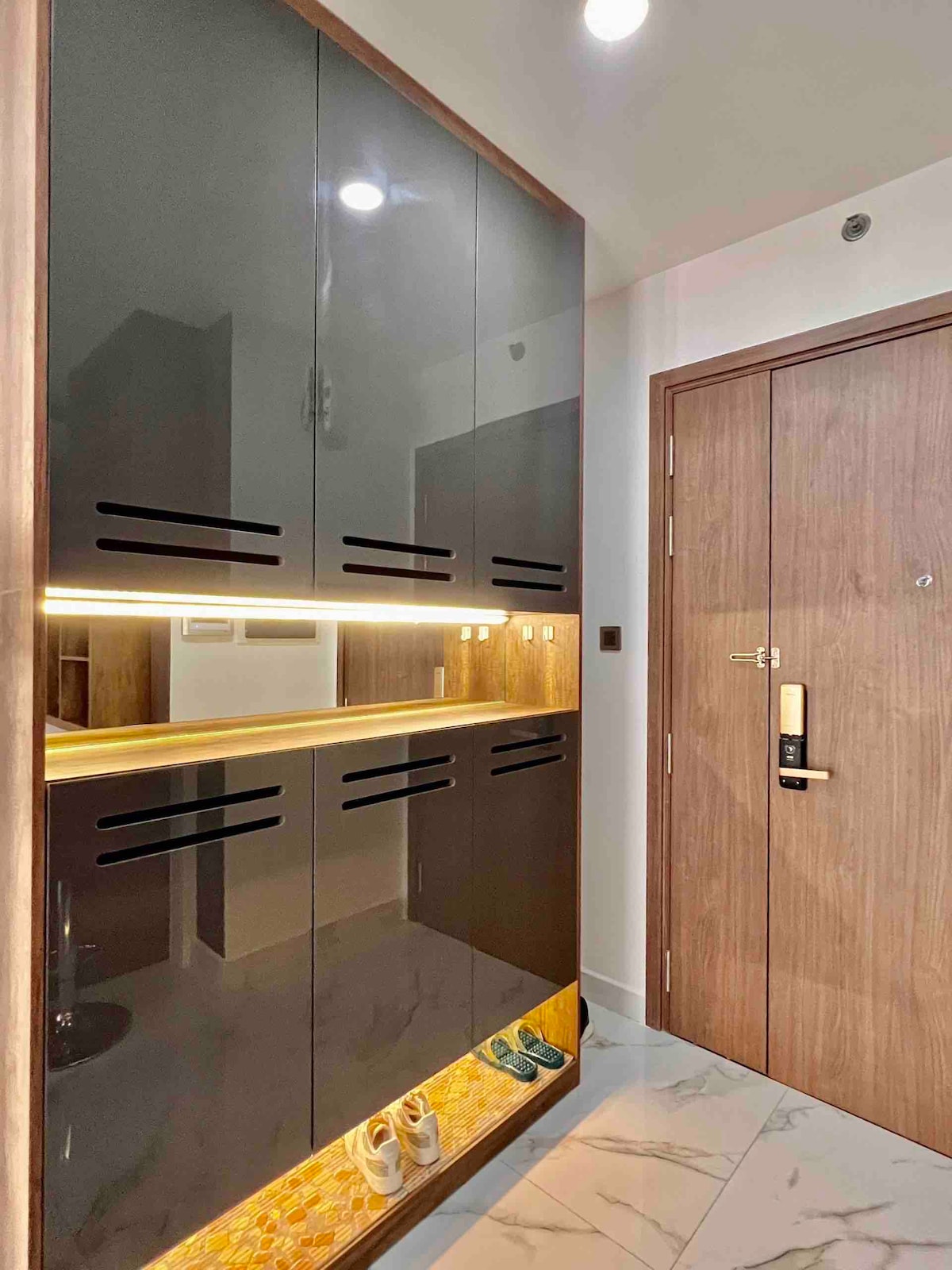 A modern entryway is presented with sleek, high-gloss cabinetry and subtle backlighting. A minimalist shoe rack displays several pairs of shoes, with the floor featuring a polished marble finish. The wooden door offers a contemporary touch, complementing the overall design.