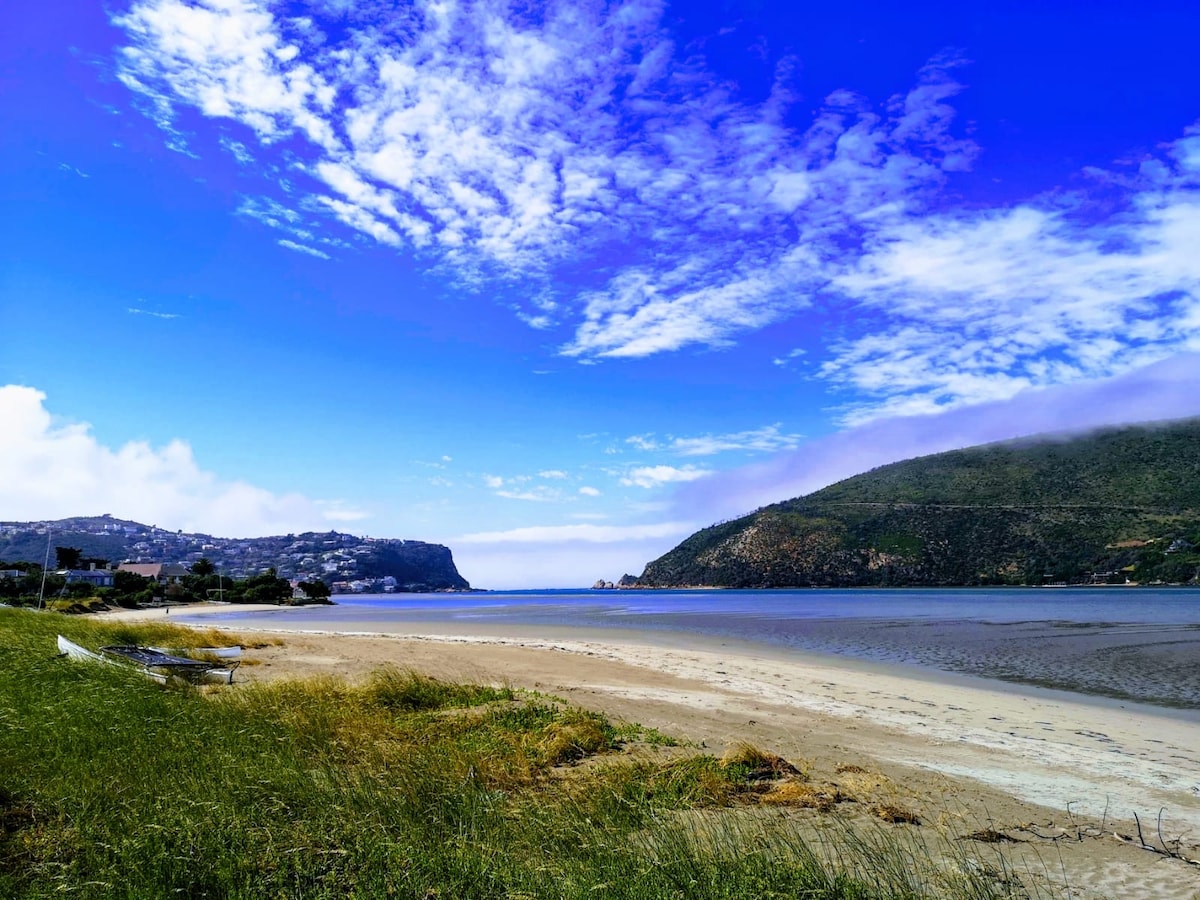 A scenic view of a beach with gentle sand leading to calm waters. Rolling hills are visible in the distance, accompanied by a vibrant blue sky filled with scattered clouds. Lush greenery borders the shoreline, enhancing the tranquil seaside setting.