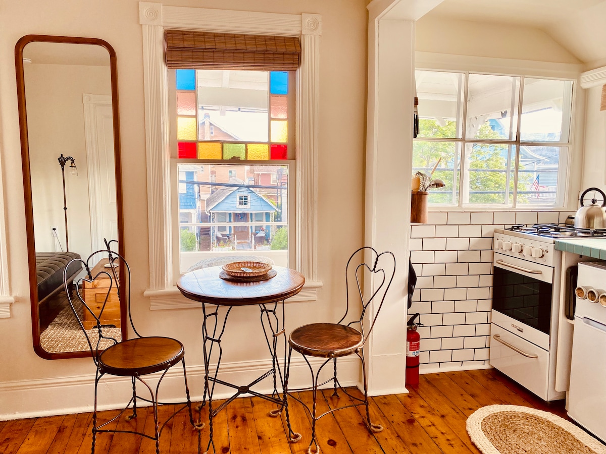 A compact dining area features a round table with two antique wrought-iron chairs, set against a backdrop of historic stained glass windows that allow light to filter through. A well-equipped kitchen is visible, showcasing white subway tiles and a classic stove.
