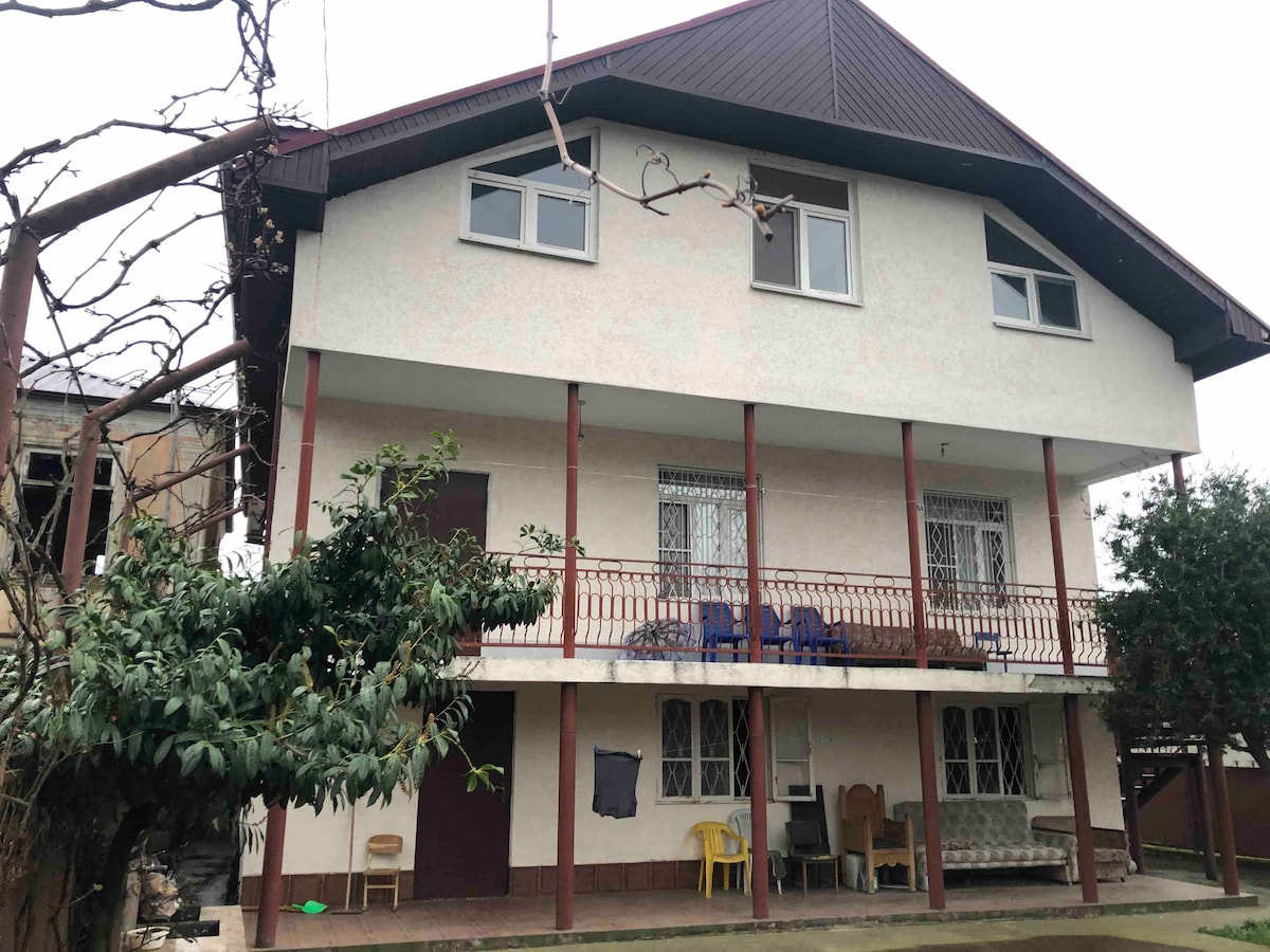 A two-story house is depicted with a simple facade and a sloping roof. A wide balcony extends across the upper level, furnished with blue chairs. The lower level features a spacious porch area with seating options. Surrounding greenery adds a natural touch to the setting.
