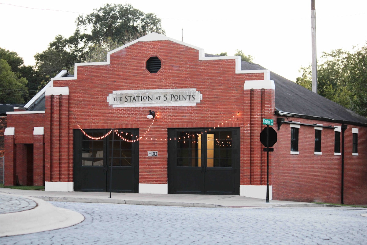 The exterior of a historic brick building is showcased, featuring large double doors and prominent signage reading 'The Station at 5 Points.' The structure is adorned with decorative string lights and surrounded by trees, with a gently curved road leading into view.