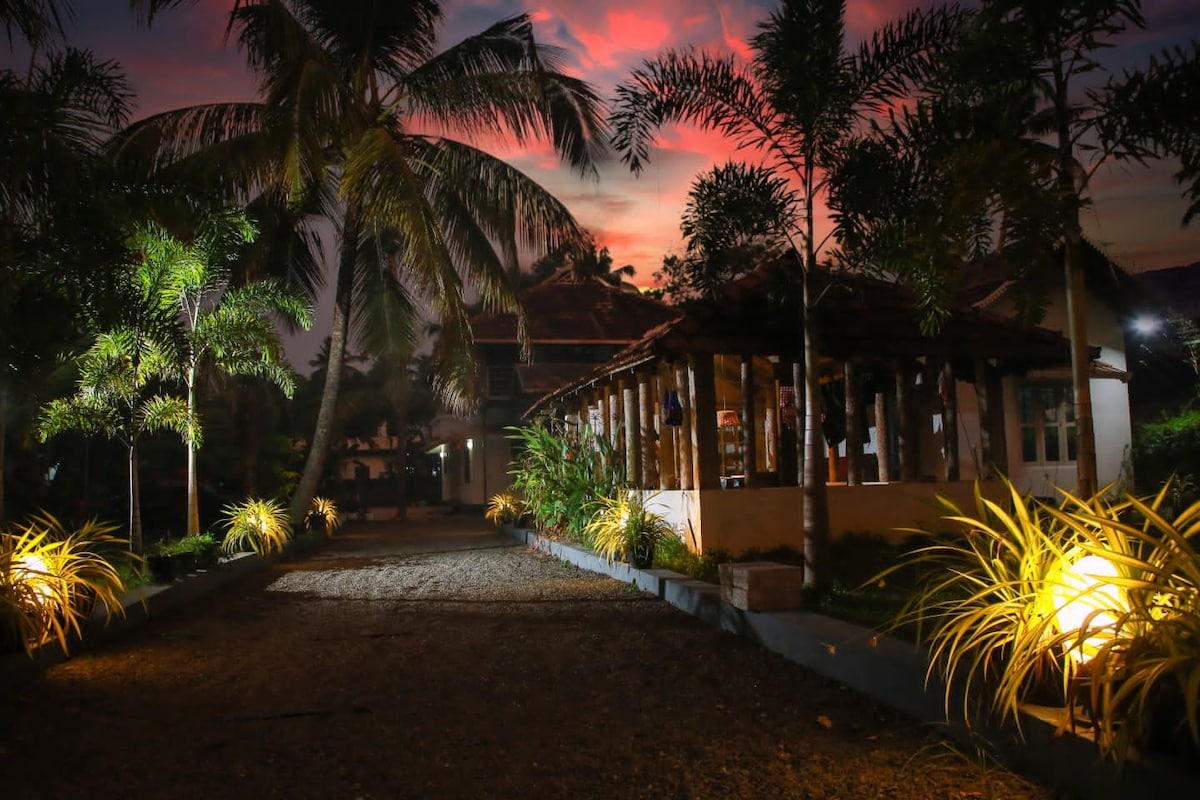 A pathway is softly illuminated by lights along the sides, leading toward a rustic structure surrounded by tropical palm trees. The sky is painted with shades of pink and orange as dusk approaches, enhancing the serene atmosphere of the setting.