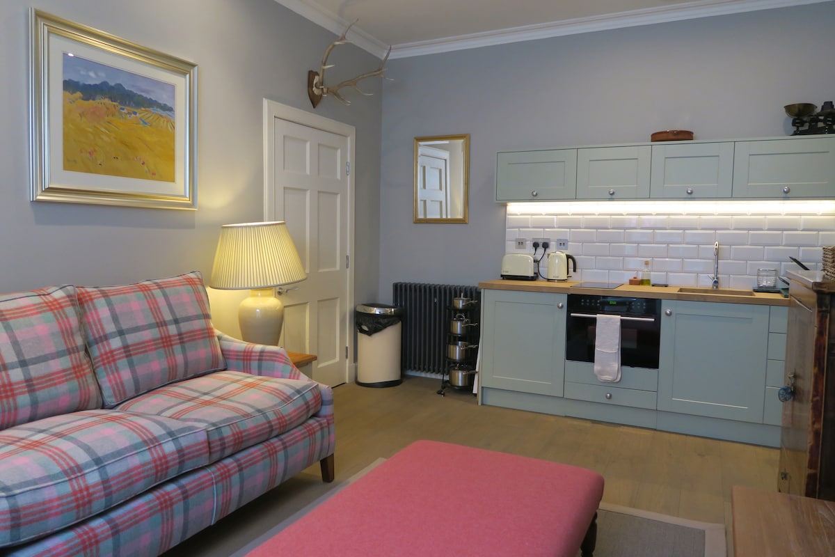 A sitting area is presented, featuring a plaid sofa and an ottoman. A kitchenette is located along the wall, equipped with basic appliances and illuminated by under-cabinet lighting. Natural light enters through a nearby window, complementing the neutral wall color and decorative wall art.