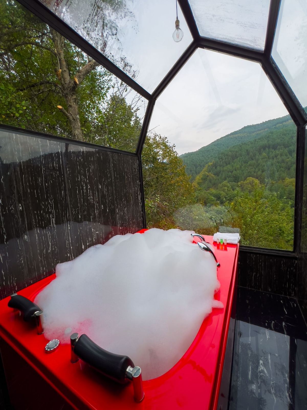 A red bathtub is filled with bubbles and surrounded by glass walls, offering views of the green mountains beyond. A natural setting is highlighted through the trees visible outside the enclosure, creating an inviting atmosphere for relaxation.