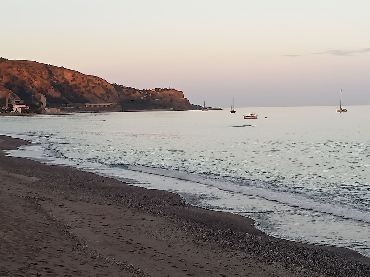 A serene coastline is depicted at sunset, with soft waves lapping against a pebbled beach. Small boats are visible on calm waters, and distant hills provide a gentle backdrop. The sky transitions from warm tones to soft blues, creating a peaceful atmosphere.