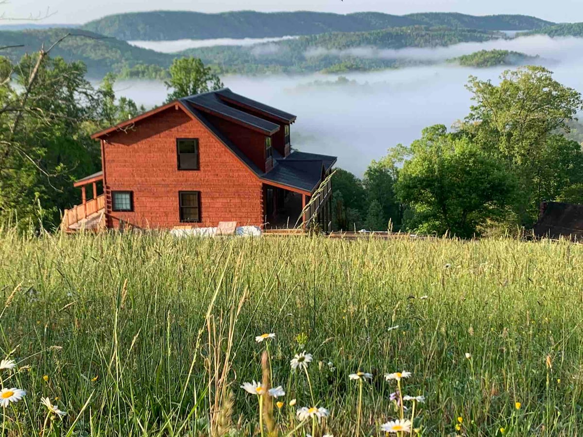 A rustic red cabin stands in a grassy field, surrounded by trees. In the background, rolling mountains are partially shrouded in mist, illustrating serene natural beauty. Wildflowers can be seen in the foreground, adding subtle color to the landscape.