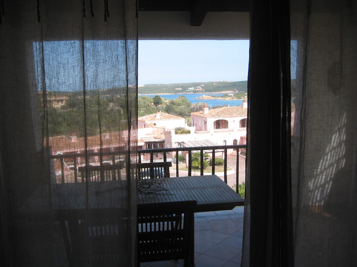 A view from the interior showcases a spacious outdoor terrace with a wooden dining table and chairs. Sheer curtains filter the sunlight, creating a soft ambiance, while the landscape reveals a glimpse of the coastline and lush greenery beyond.