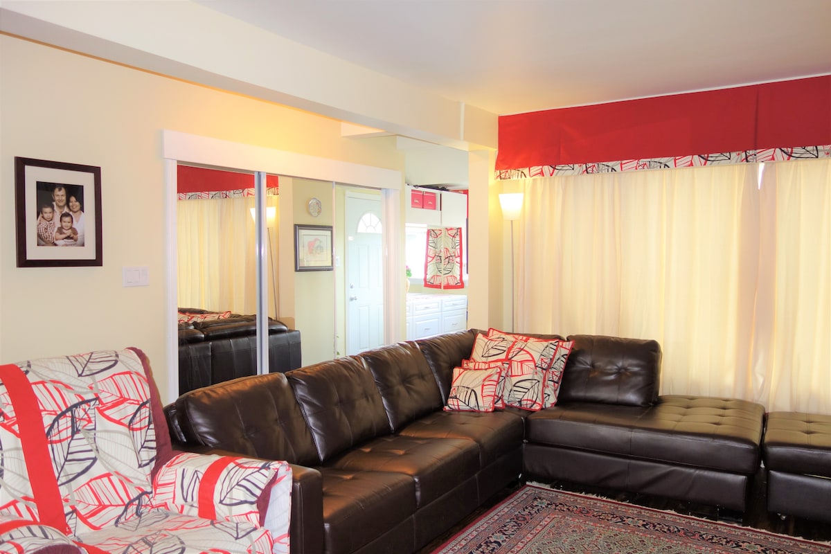 A modern living area features a stylish, dark leather sectional sofa adorned with a vibrant patterned throw pillow. A light-colored wall provides a neutral backdrop, while large windows draped with sheer curtains invite natural light. A mirrored panel reflects the room, enhancing the spacious feel.