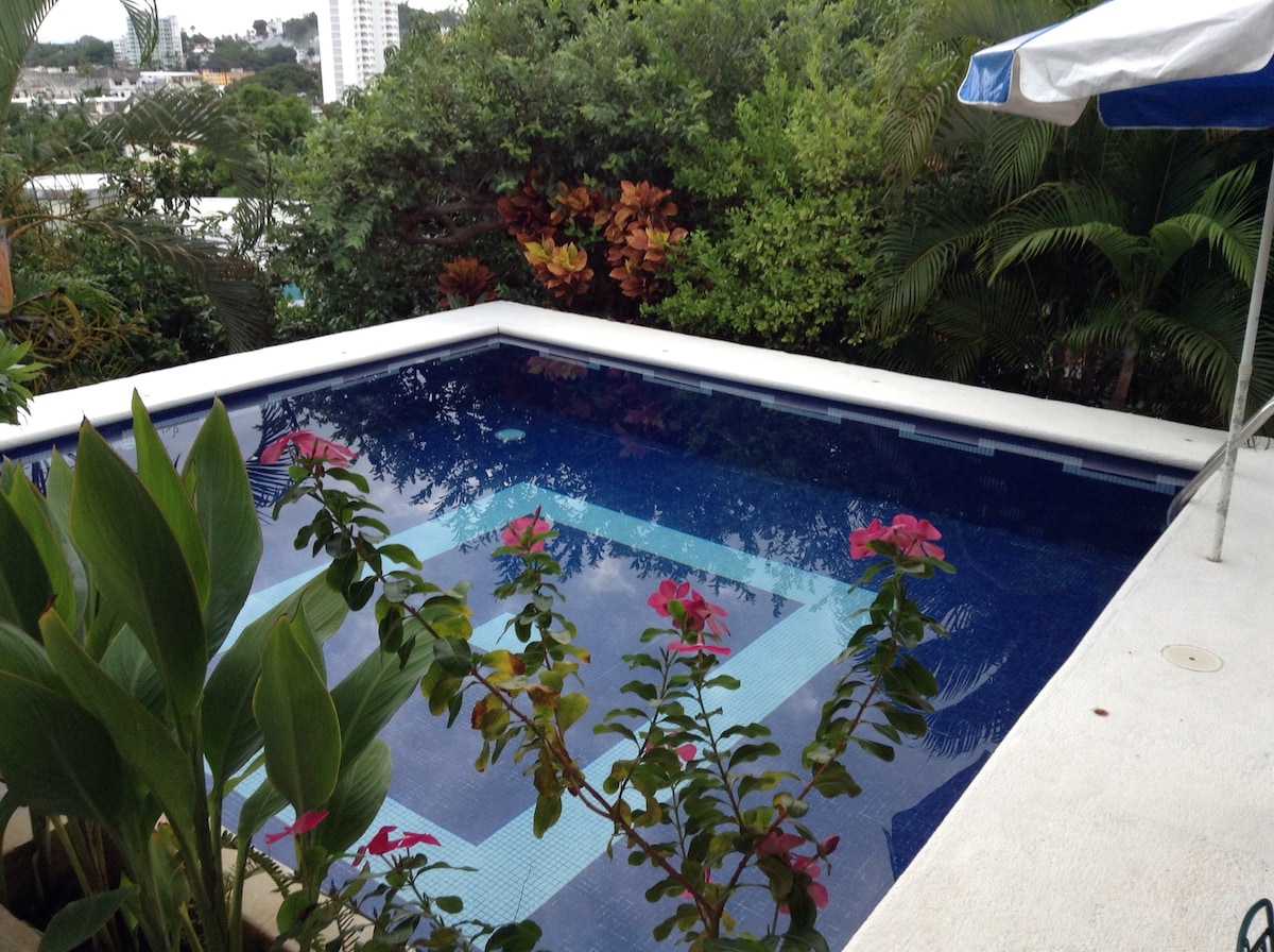 A serene swimming pool is framed by lush greenery and colorful flowering plants. The water appears calm, reflecting the surrounding foliage and providing a peaceful atmosphere. An umbrella is visible at the edge, offering shade in the sunny environment.