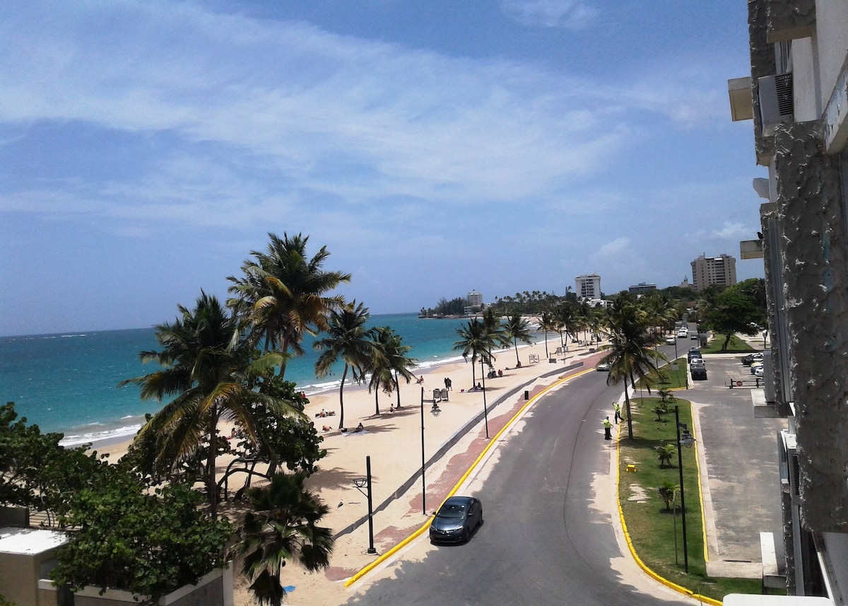 A scenic view captures the sandy beach lined with palm trees and the turquoise ocean under a clear blue sky. The waterfront road curves gently, leading towards the horizon, where buildings can be seen in the distance.
