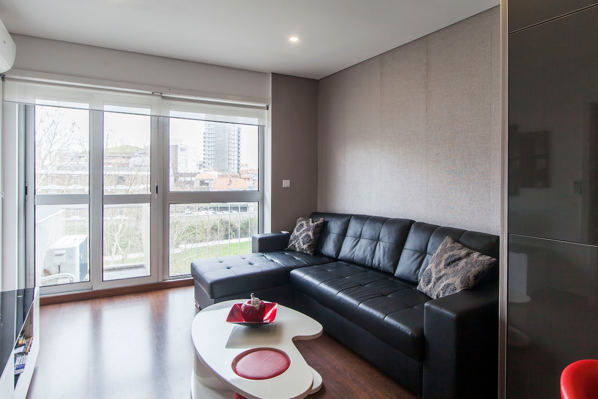 A modern living area is featured, showcasing a large black sectional sofa and a white coffee table with a decorative bowl. Natural light flows through the sliding glass door, which leads to a view of the outdoors, enhancing the open and airy feel of the space.