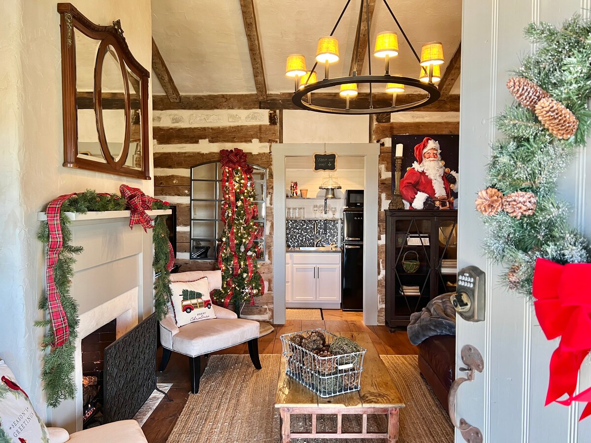 The entryway of the Dr. Okerholm Loghaus features a warm and inviting interior with exposed wooden beams. A large mirror hangs above a decorative fireplace, surrounded by festive garlands. A plush sofa and armchair are positioned around a coffee table, with seasonal decor accentuating the space.