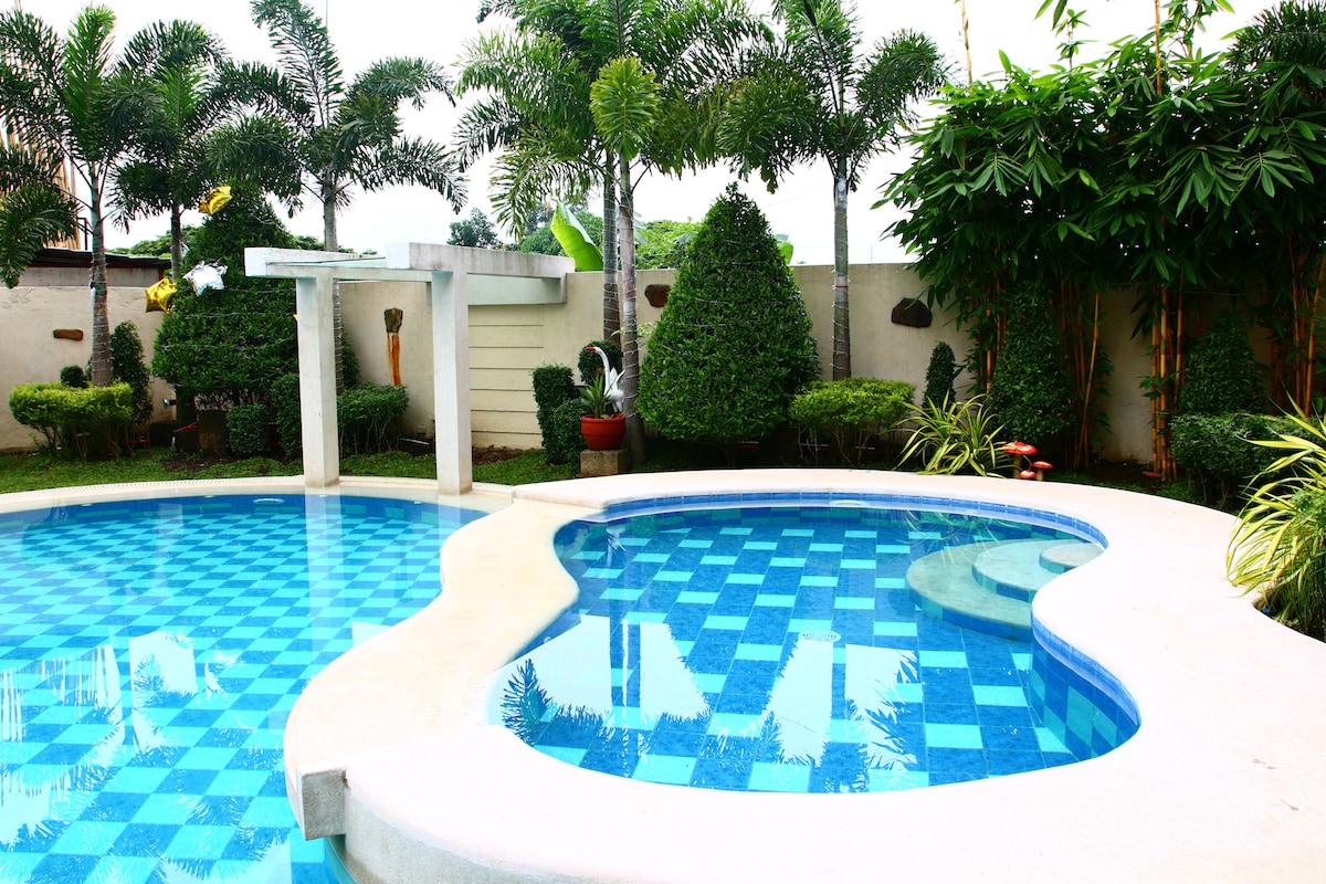 The outdoor swimming pool features a unique design with checkerboard patterns in shades of blue. A glass waterfall is visible at one end, surrounded by tropical plants and neatly trimmed hedges. The area offers a serene and inviting environment for relaxation.