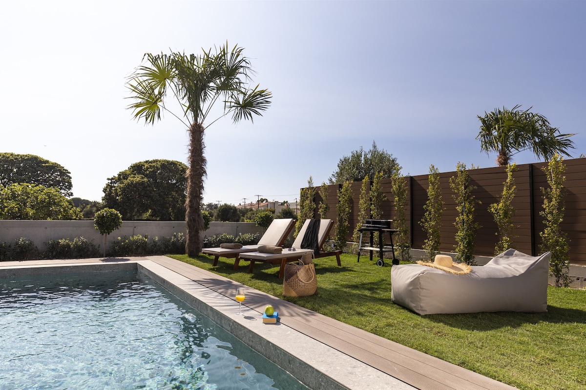 A serene outdoor area features a private pool surrounded by lush greenery. Two sun loungers are positioned beside the pool, while a small table and grill are visible in the background. A cozy seating option is also present, providing a relaxed atmosphere.