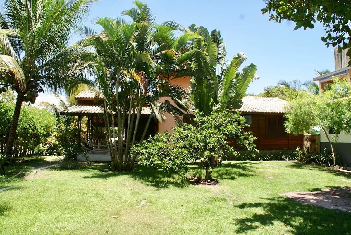 A lush green lawn is surrounded by tropical plants and palm trees, providing shade and a natural atmosphere. A wooden house with a thatched roof can be seen in the background, while a spacious veranda invites relaxation in the serene outdoor setting.