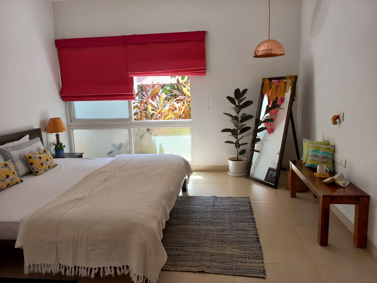 A bright bedroom features a spacious bed adorned with neutral bedding and decorative pillows. Natural light enters through a large window, illuminating a cozy seating area. A potted plant and a floor mirror add an inviting touch, while a wooden table holds a small decorative arrangement.