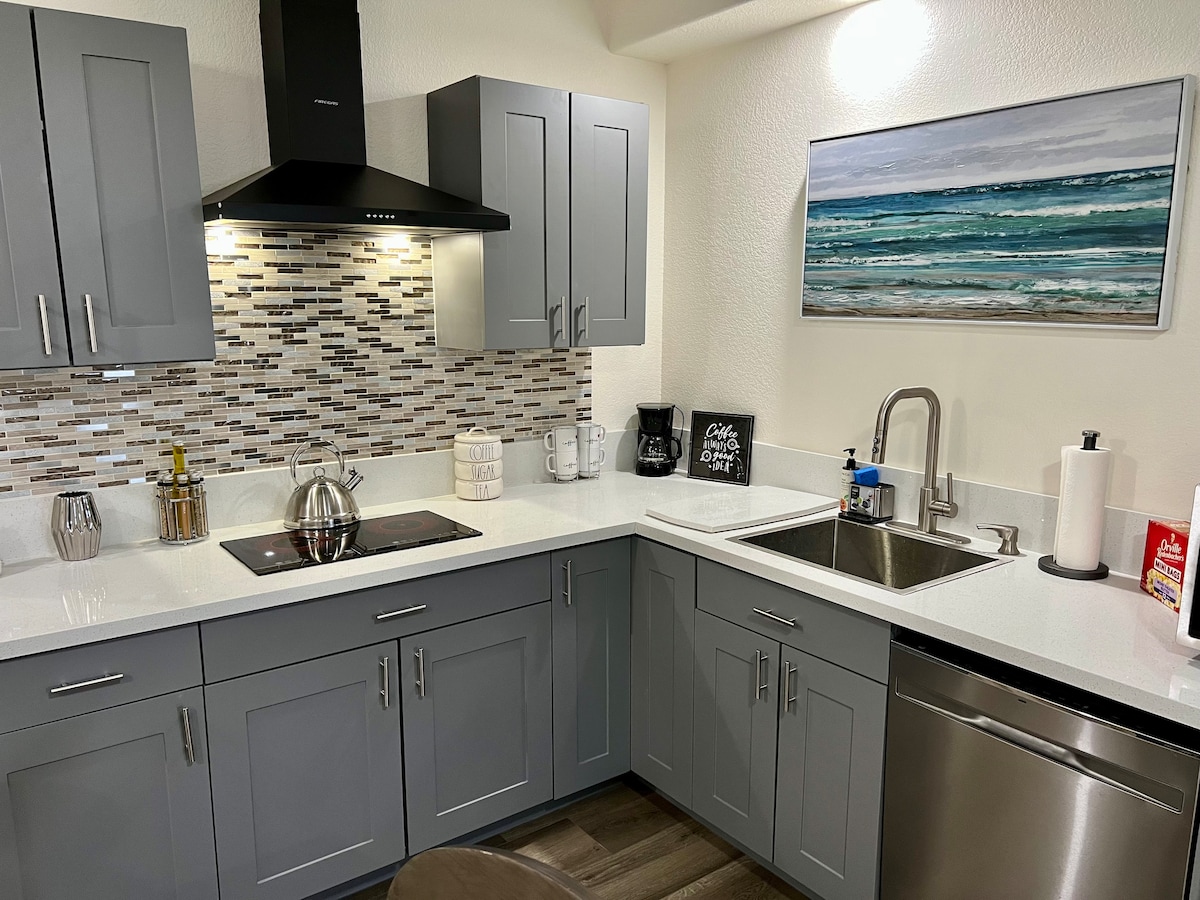 A modern kitchen featuring gray cabinetry and a sleek design. A minimalist stove hood is mounted above the stovetop, while the countertops display gleaming materials. A double sink and a dishwasher are included, alongside decorative tile backsplash, offering a clean and functional space.