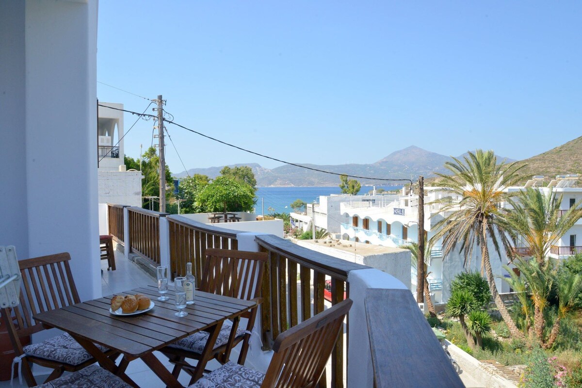 A spacious balcony is featured with a wooden table and chairs, inviting relaxation with views of the sea and surrounding landscape. Green palm trees and white buildings are visible in the background, complemented by clear blue skies and distant mountains.