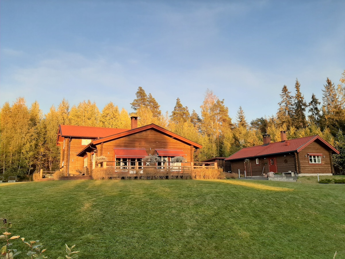 The exterior of a wooden guest cottage is shown, surrounded by lush greenery and autumn foliage. A spacious deck extends from the main building, which features large windows. A separate smaller building is visible beside the main structure, both topped with red roofs.