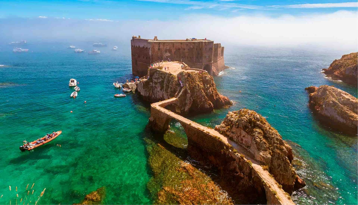 An aerial view captures a rugged coastline featuring a historic stone fort on an island. The fort is surrounded by clear blue water, dotted with small boats. A narrow stone pathway connects the fort to the mainland, highlighting the scenic seascape and rocky outcrops.