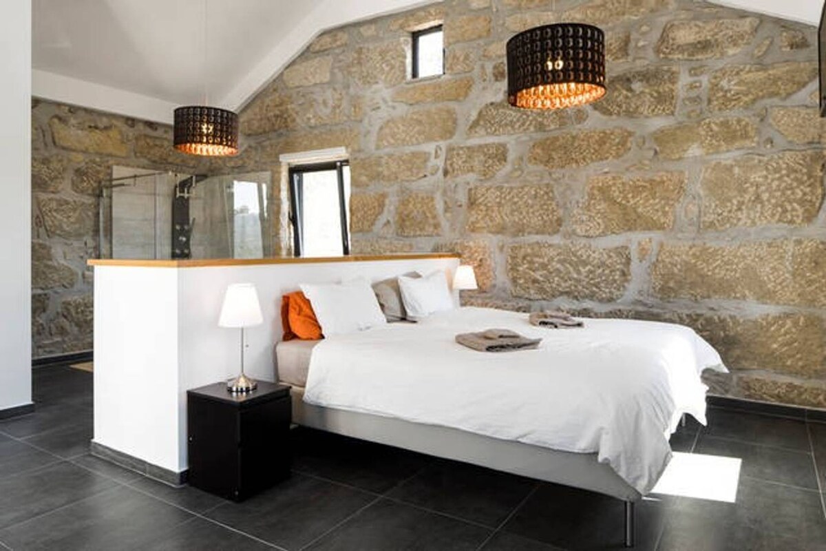 A spacious sleeping area features two twin beds dressed in crisp white linens. Soft lighting is provided by two decorative pendant lights above, complementing the natural stone walls. A nightstand with a lamp sits beside one bed, enhancing the overall calming aesthetic.