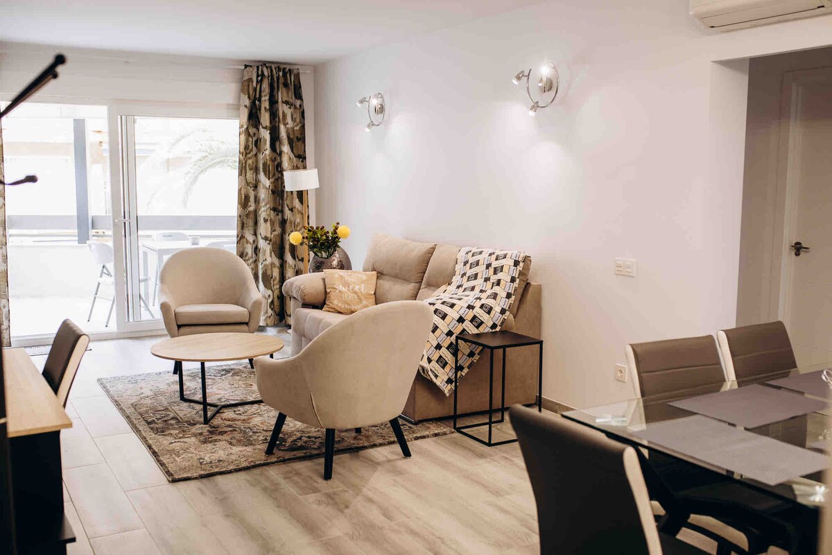 A modern living area features a beige sofa adorned with decorative cushions and a patterned throw. Two light-colored armchairs are positioned beside a round coffee table, while a dining set with dark chairs is visible. Natural light fills the space through large sliding doors.