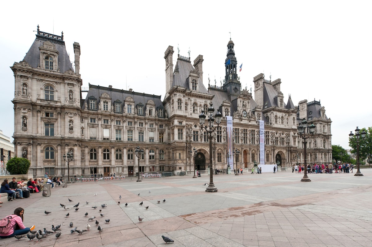A grand historic building features intricate architectural details and a prominent clock tower. The surrounding area is paved, with scattered pigeons on the ground. Visitors can be seen in the background, adding to the bustling atmosphere of this iconic public square.