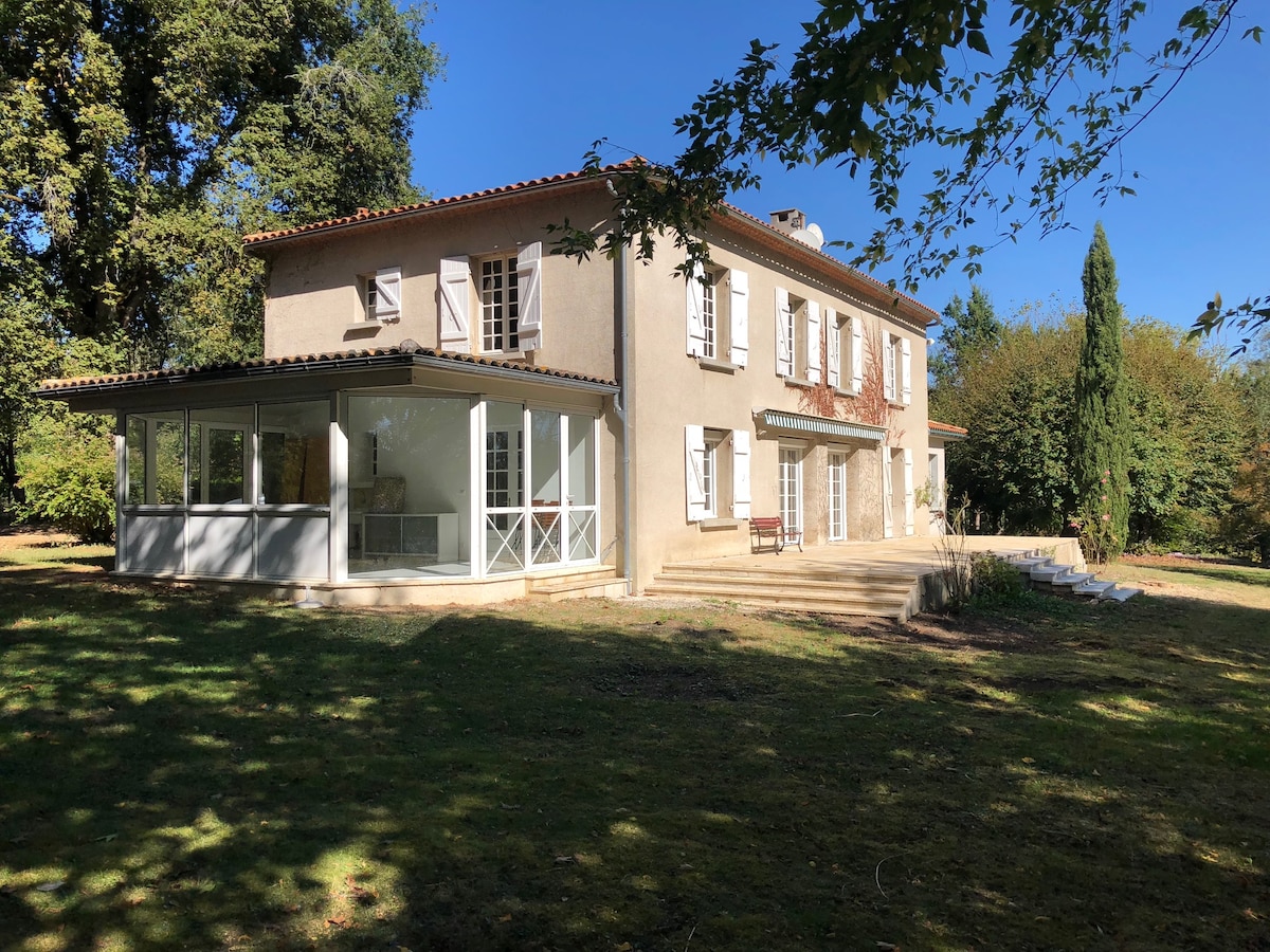 The house is a two-story structure with a light-colored facade and traditional shutters. A spacious veranda with large windows overlooks the garden, which is surrounded by trees. Steps lead from the veranda to the lawn area, creating a seamless connection between indoor and outdoor spaces.