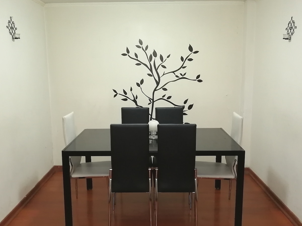 A dining area is presented with a black table surrounded by six modern chairs. A decorative wall decal of a tree branch enhances the ambiance. Natural light brightens the wooden floor, and two wall-mounted sconces provide additional illumination.