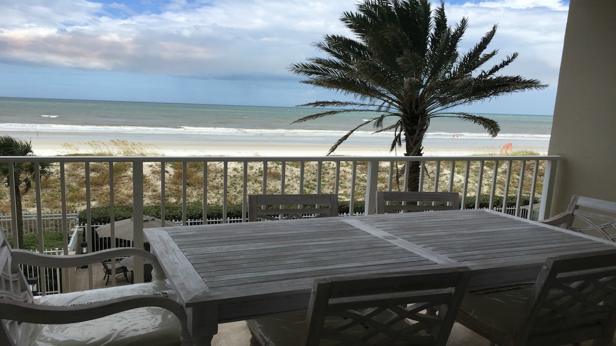A spacious balcony features a wooden dining table accompanied by multiple chairs, overlooking a sandy beach and the ocean. Palms sway gently in the breeze, with a clear view of the sea and distant shoreline.