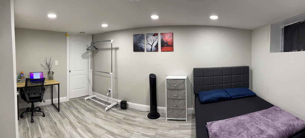 A well-lit room contains a spacious bed with blue pillows and a light grey headboard. A work desk with a monitor sits adjacent, accompanied by a chair. A clothing rack is positioned beside a tall dresser, while art pieces featuring trees adorn the wall.