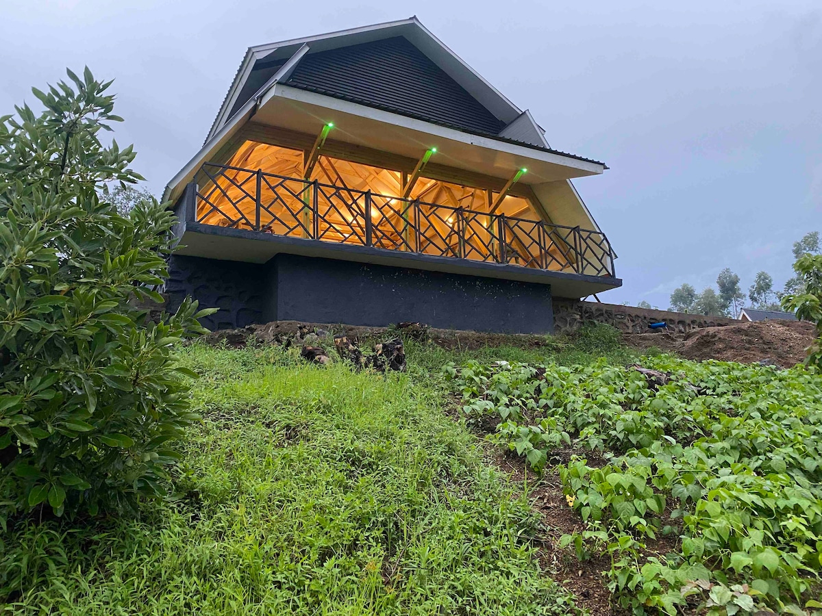 The modern structure is set against a lush green hillside, featuring a spacious balcony with wooden railings. Soft lighting illuminates the interior from large glass doors, creating an inviting entrance under a cloudy sky.