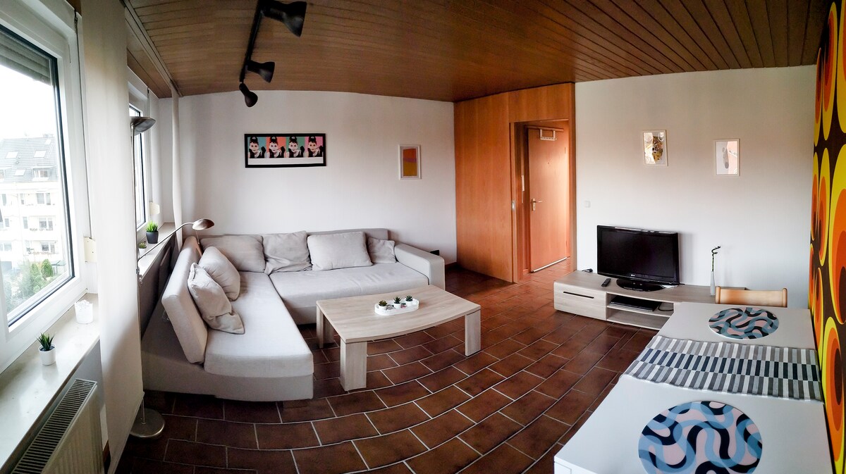 A bright living room features a spacious L-shaped couch, paired with a simple wooden coffee table. A vibrant wall art piece adds character, while a modest TV unit is positioned against the wall. Natural light comes through large windows with minimal decorations.