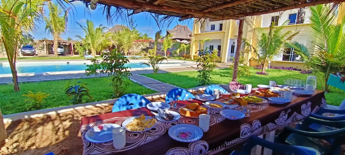 A dining table is set under a shaded area, featuring a variety of breakfast items arranged on plates. In the background, a swimming pool is visible, surrounded by palm trees and lush greenery, creating a tranquil outdoor setting.