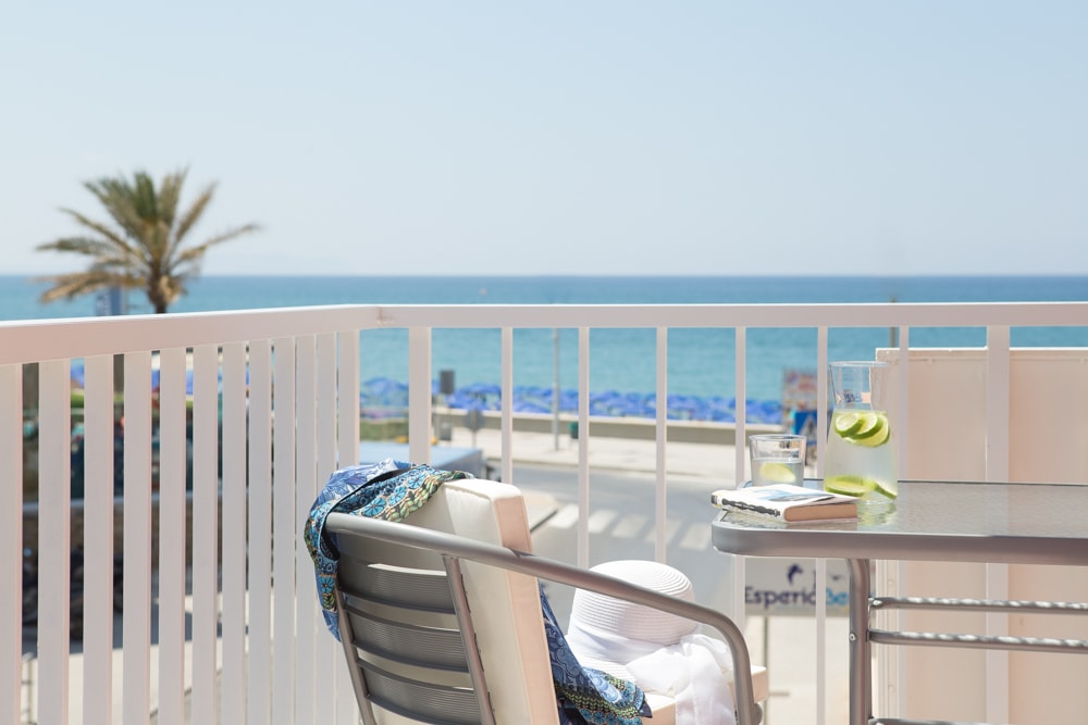 A balcony offers a view of the beach and sea, featuring a table with a refreshing drink and a folded beach towel. A palm tree is visible in the distance, and sun loungers are arranged on the sandy shore, enhancing the coastal atmosphere.