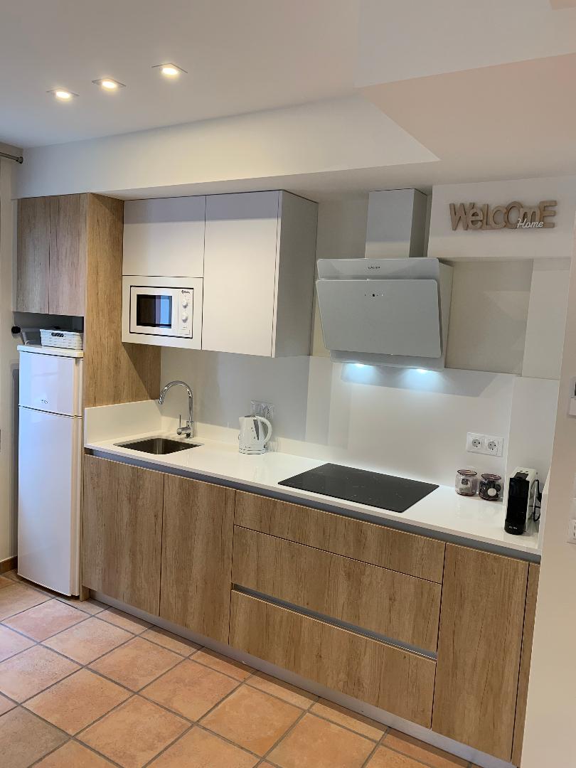 A modern kitchen features a combination of light wood and white cabinetry. A sleek microwave and a white kettle are visible on the countertop, alongside a stovetop. The sink is positioned beneath a wall-mounted extractor hood, and a small refrigerator stands nearby.