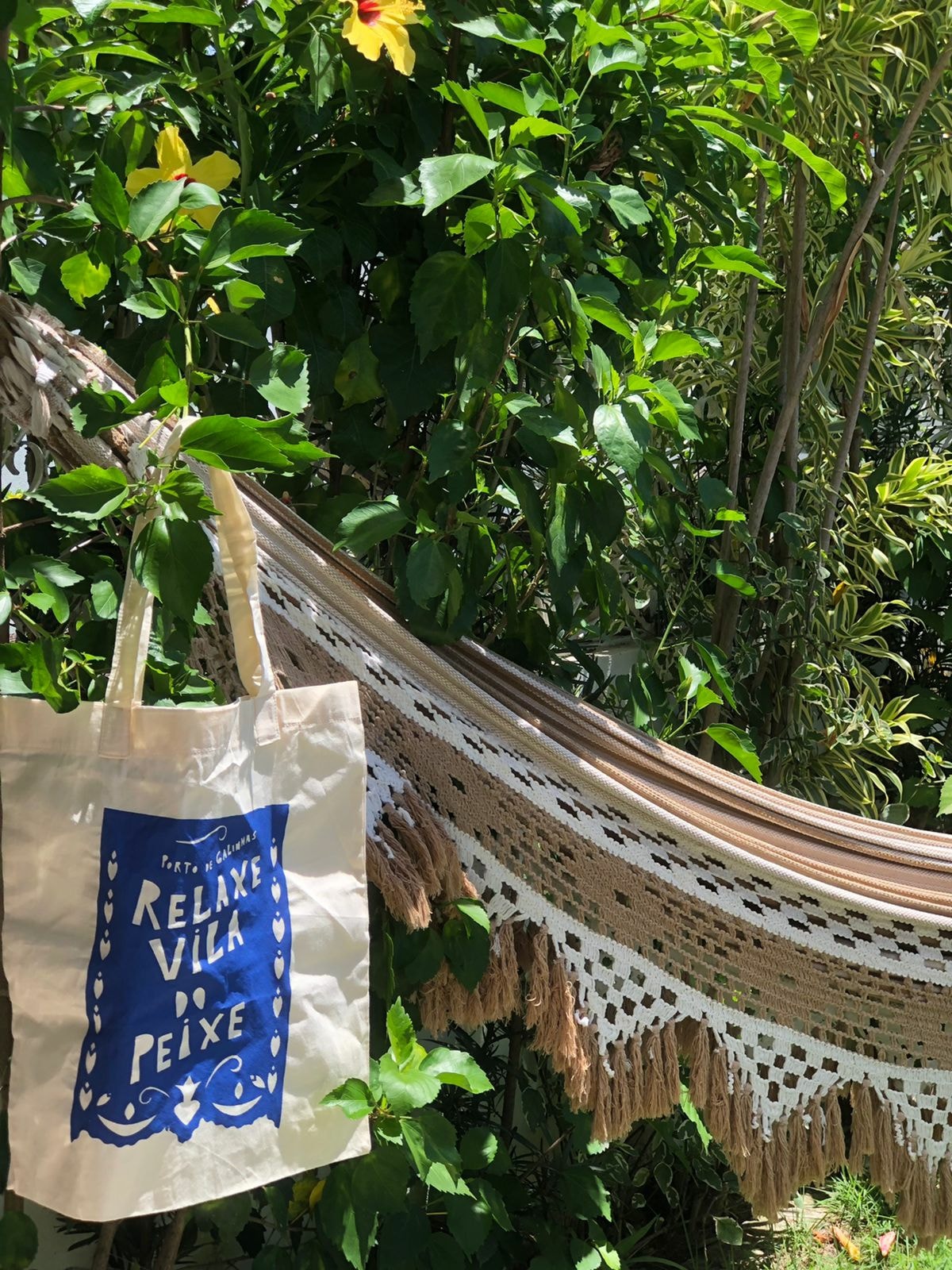 A woven hammock is draped between lush green foliage, inviting relaxation. A fabric tote bag hangs nearby, featuring a decorative design that reads 'Relax Vila Peixe.' The vibrant greenery enhances the serene atmosphere of the garden space.
