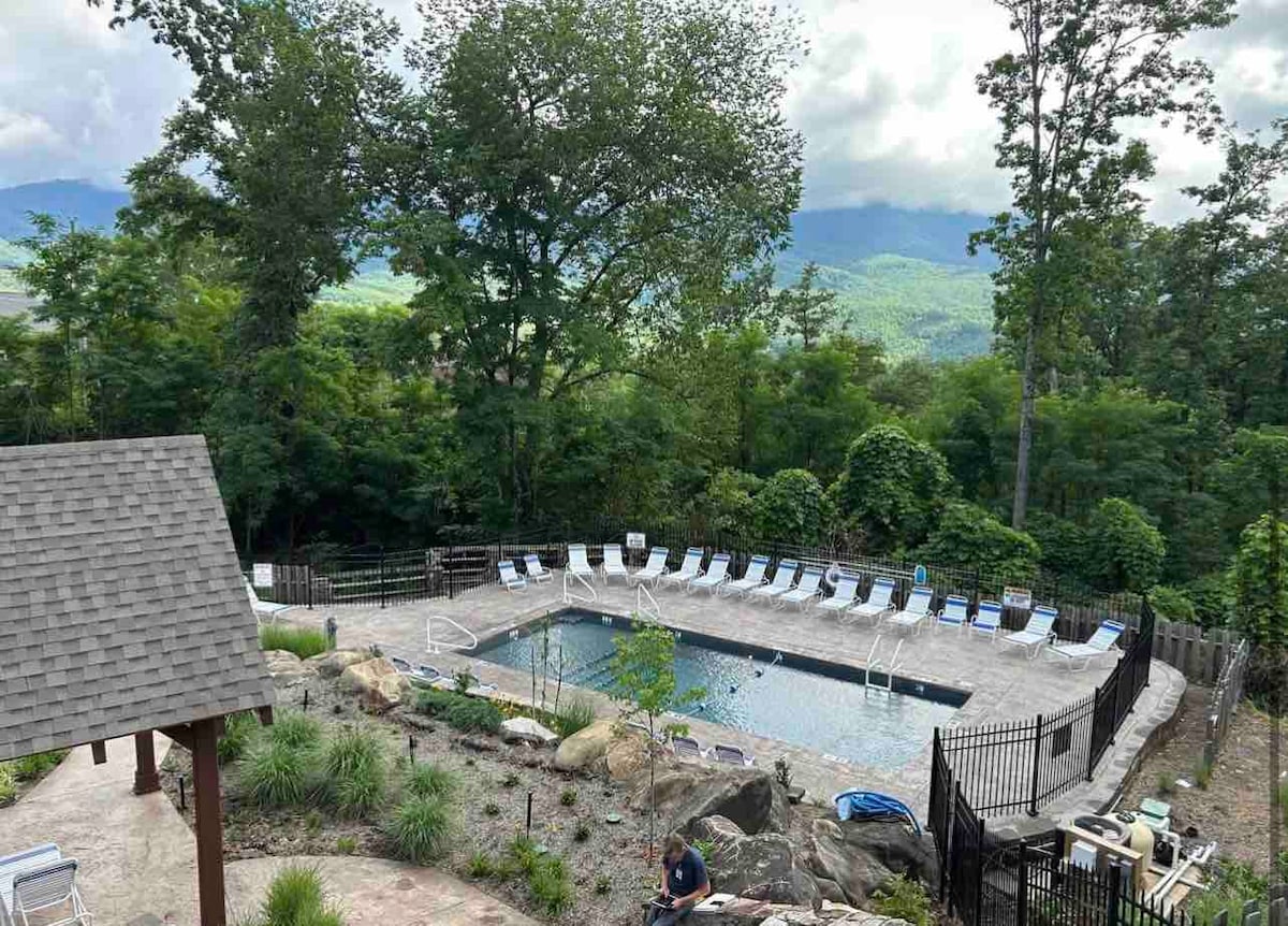 An outdoor pool area is surrounded by greenery and features lounge chairs arranged along the deck. The scenic backdrop reveals rolling hills and mountains, creating a serene atmosphere. A small gazebo is partially visible beside the pool, enhancing the tranquil outdoor setting.