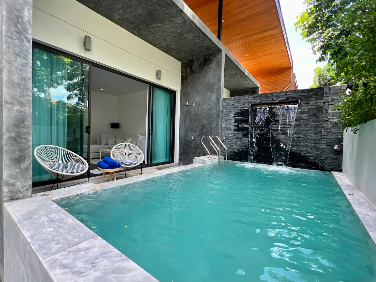 A private pool area features modern stone walls and a cascading water feature, creating a serene outdoor space. Two stylish chairs are positioned beside the clear water, which reflects natural light from the surrounding greenery.