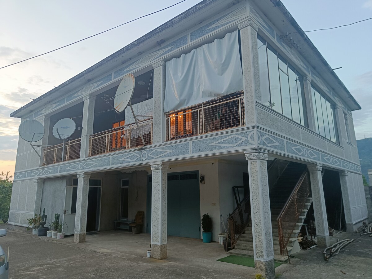 A two-story building is depicted, featuring large glass windows covered with white fabric curtains. Satellite dishes are affixed to the exterior walls. A covered entrance provides access to the upper level, while the ground floor showcases a spacious porch area, surrounded by greenery.