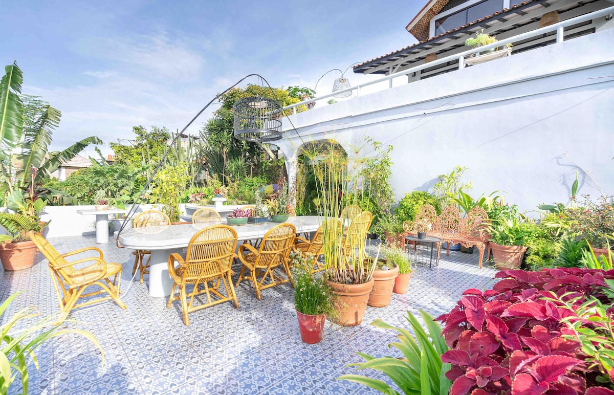 A spacious outdoor terrace features a large marble dining table surrounded by wicker chairs, inviting gatherings. Lush greenery and potted plants create a vibrant atmosphere, while the intricate blue tile flooring adds character to the area. Sunlight filters through the surrounding foliage.