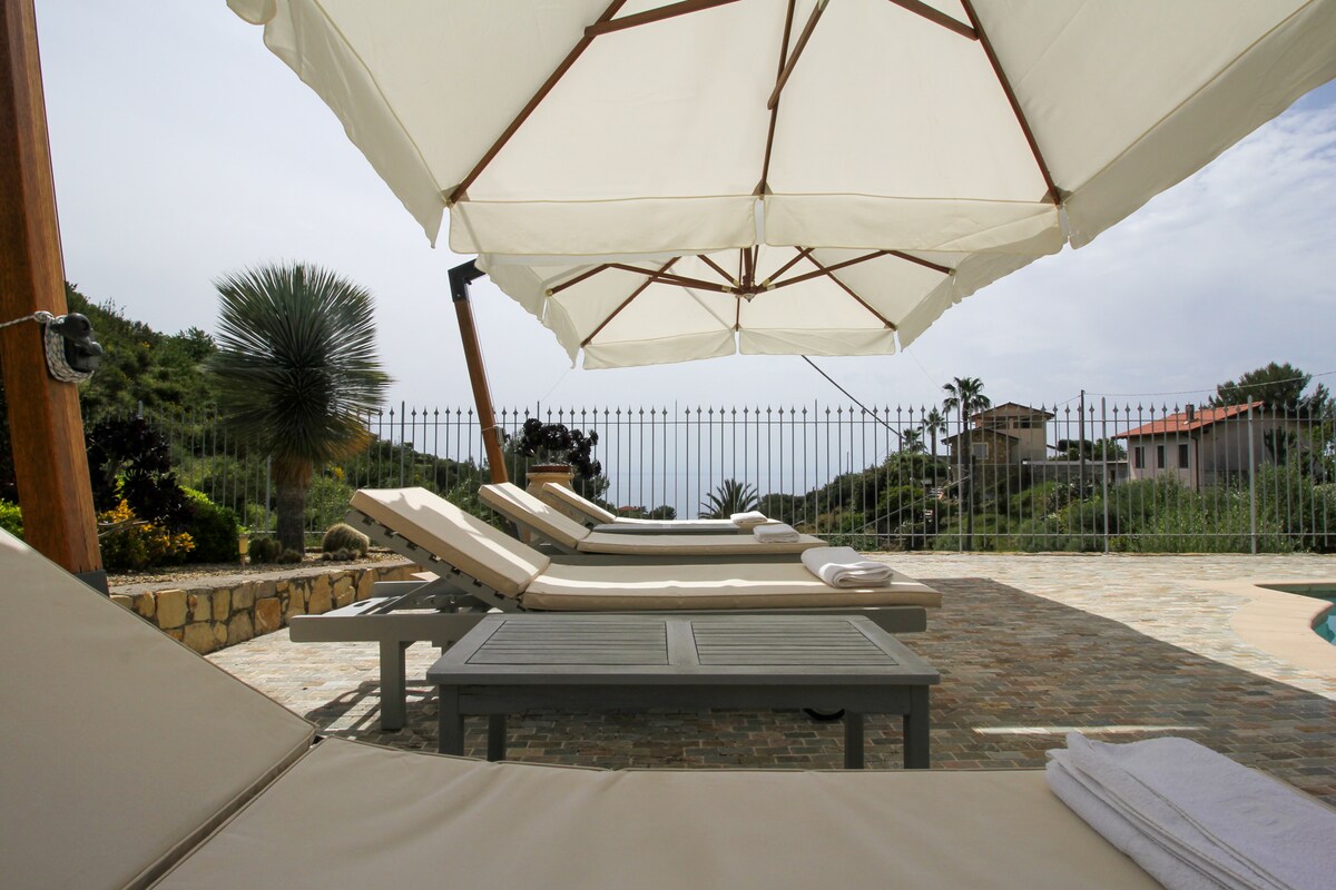 The image captures a tranquil poolside area featuring several lounge chairs arranged under large umbrellas. The space is surrounded by lush greenery, with a view of distant villas and palm trees in the background. A soft towel rests on one of the lounge chairs.