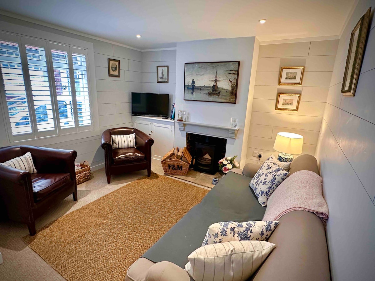 A cozy lounge area features a comfortable sofa adorned with patterned cushions. Two leather armchairs are positioned nearby, facing a television and a log-burning fireplace. Soft lighting and a jute rug complement the wooden decor, creating a welcoming space for relaxation.