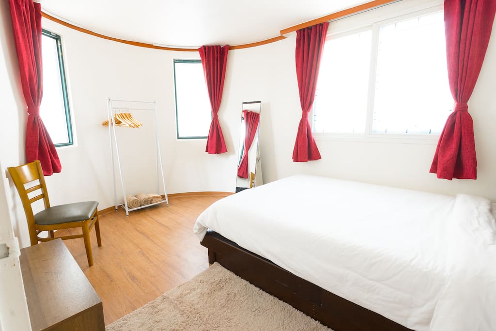 This middle bedroom has a unique semi circle design. With a queen bed, mirror, hanger, night stand, chair and a shelf with Seoul information booklets.