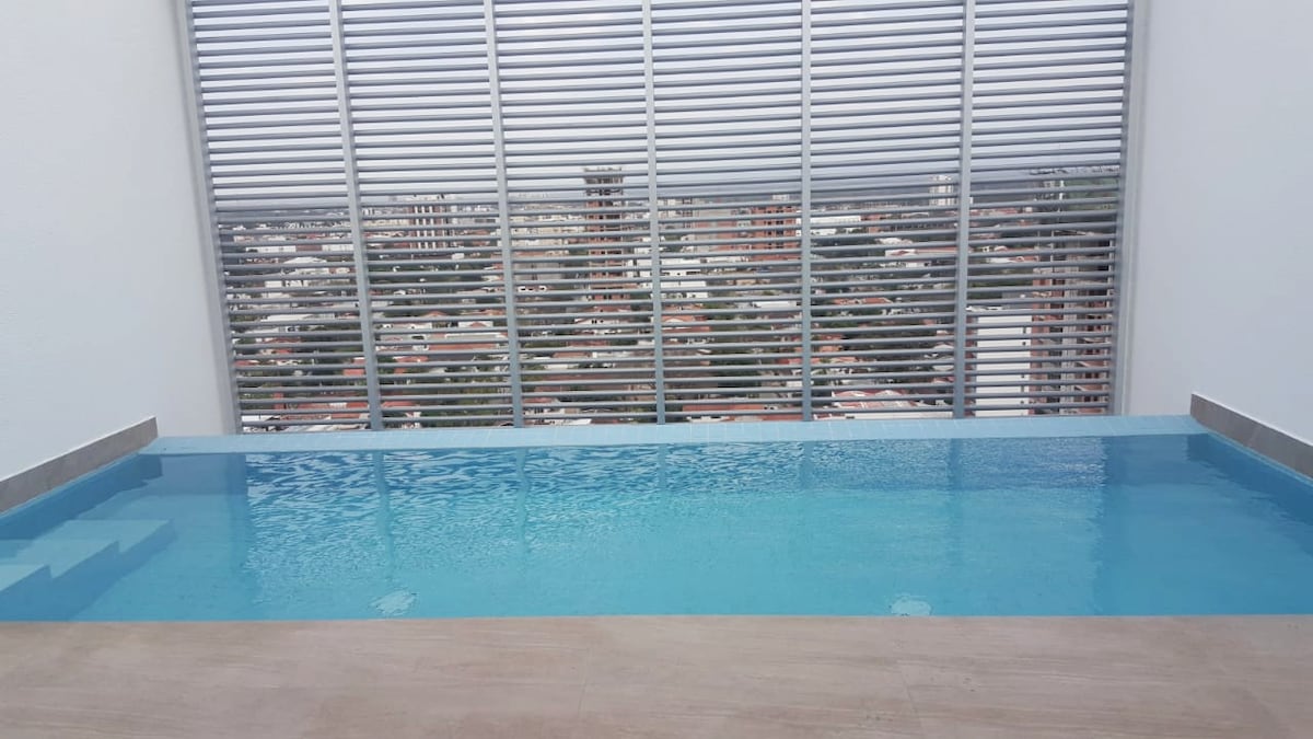 A serene swimming pool is visible, featuring clear blue water reflecting natural light. The pool overlooks a cityscape behind horizontal window blinds, creating a sense of privacy while allowing for scenic views. The surrounding area is minimally designed for a clean aesthetic.